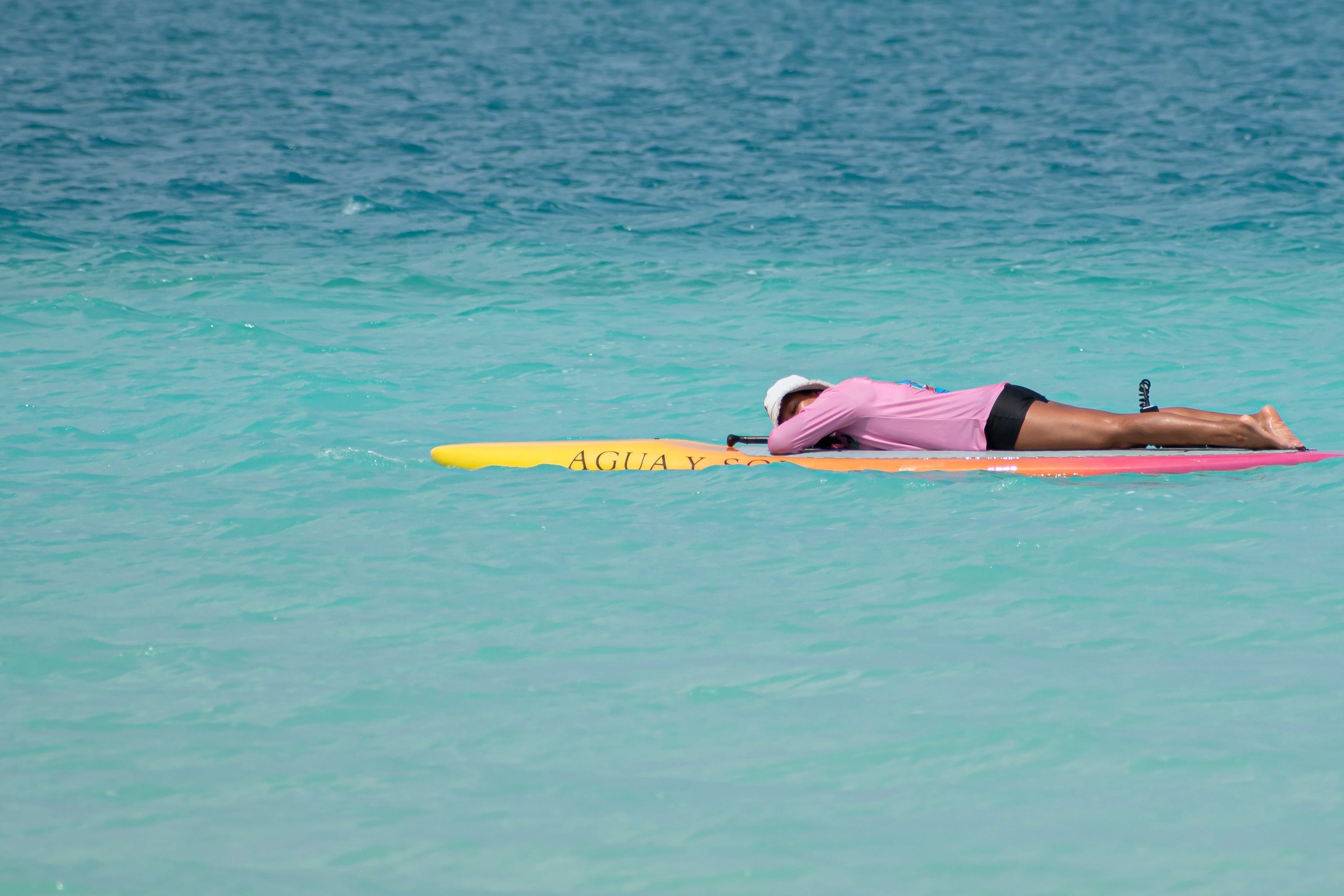 A person lies prone on a yellow and pink surfboard floating in clear turquoise ocean water, wearing a pink  upf 50 shirt and black shorts.